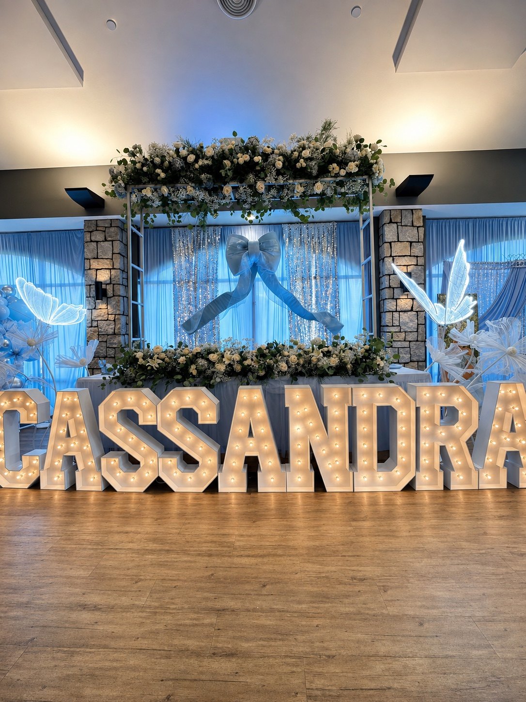 Cassandra’s XV celebration with blue and white floral decor, glowing butterflies and illuminated CASSANDRA marquee letters