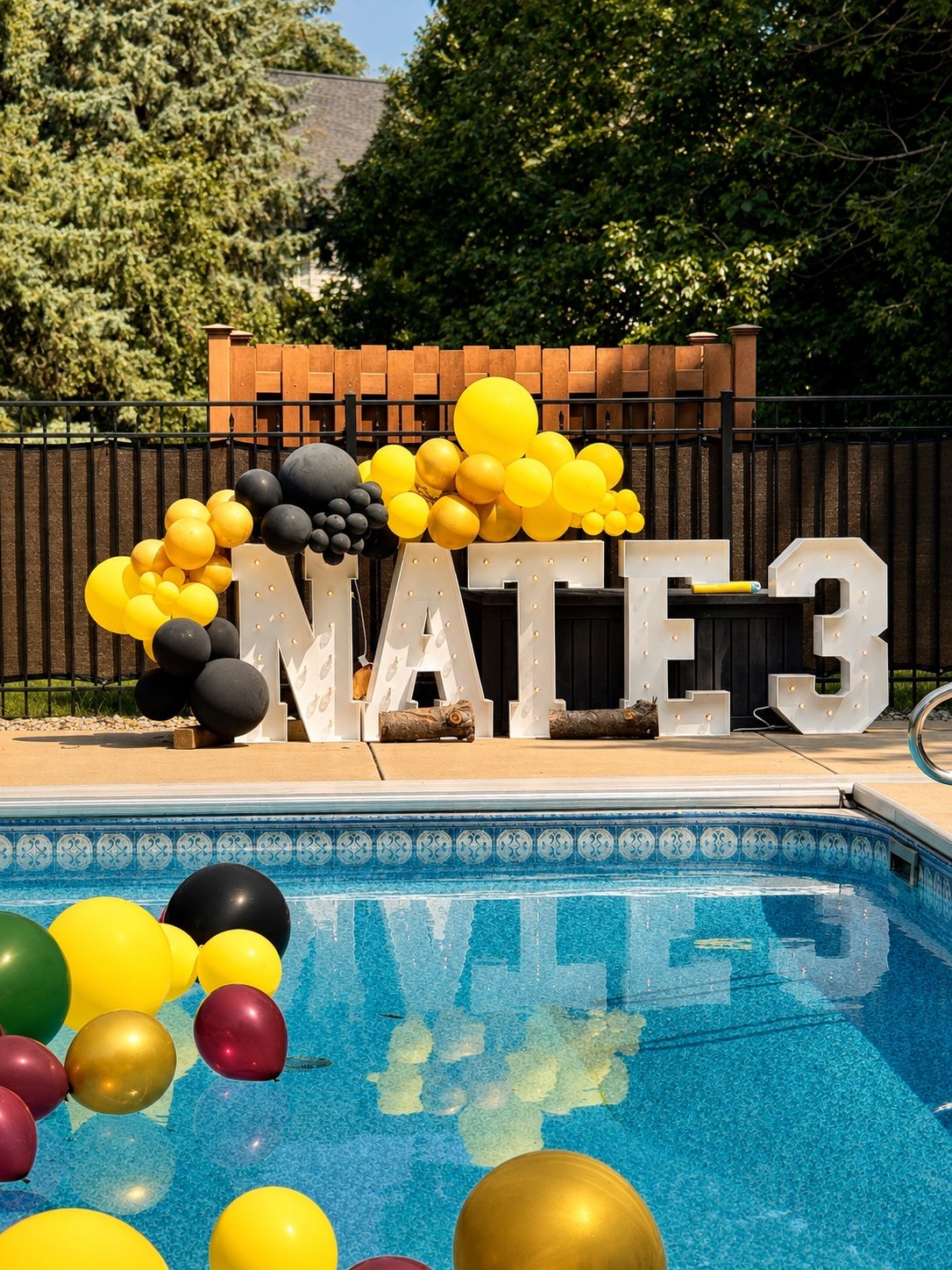 Illuminated NATE 3 letters beside a pool with yellow and black balloons