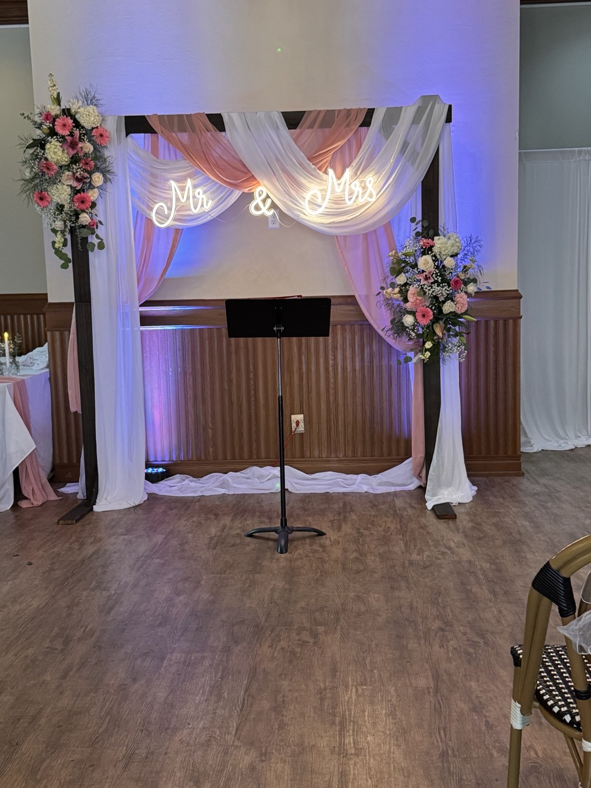 White and blush draped ceremony arch with flowers and an illuminated Mr and Mrs sign