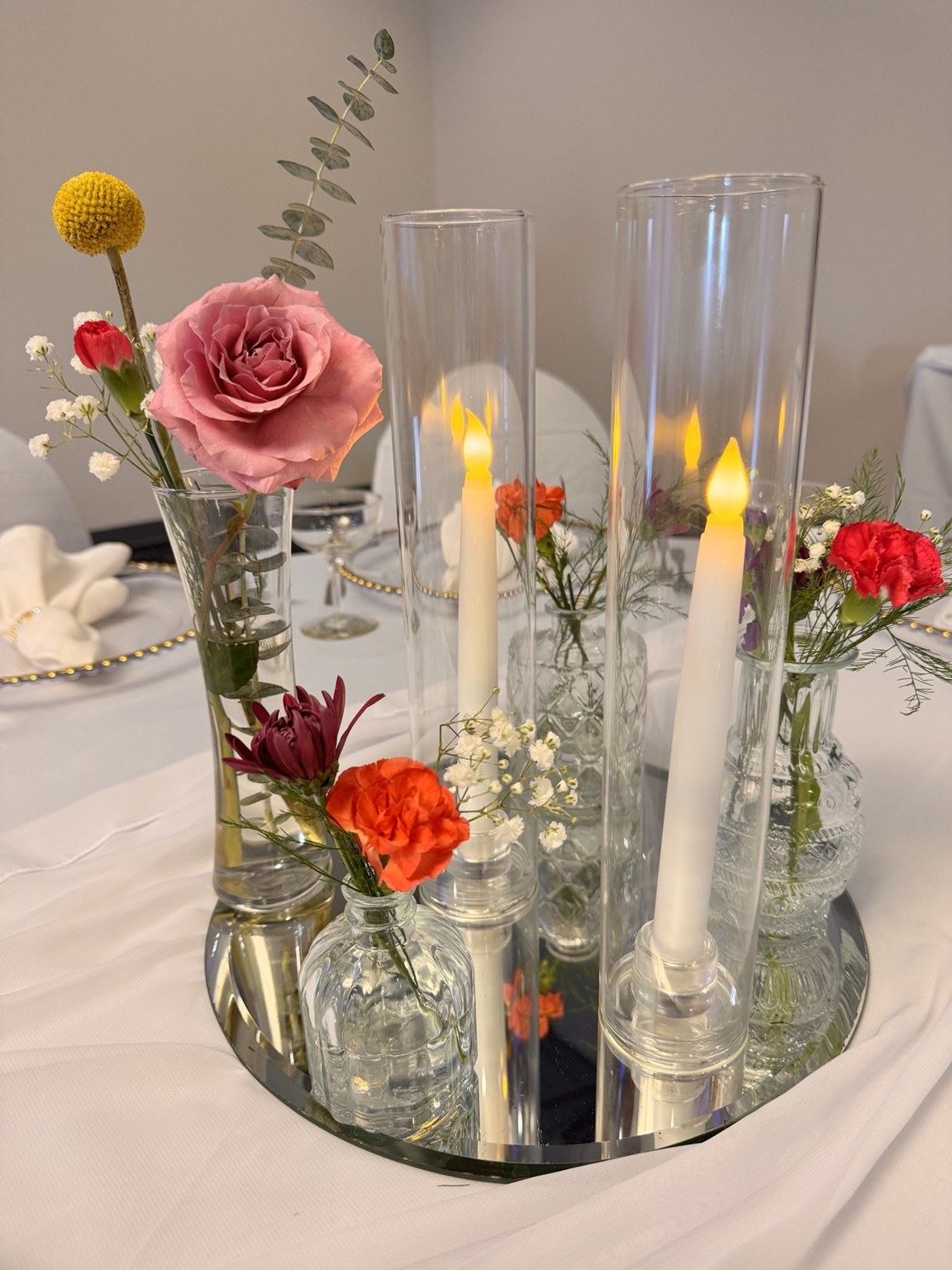 Bud vases and glowing candles arranged on a mirror with colorful flowers