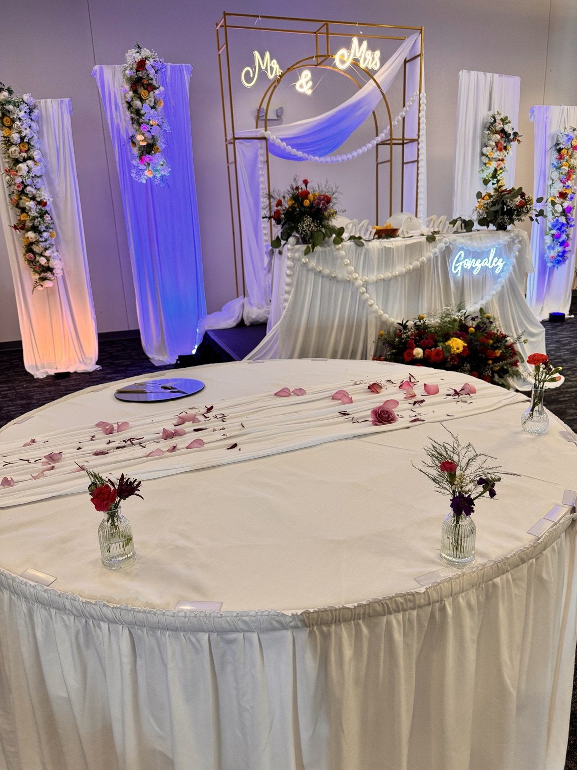 White sweetheart table with scattered rose petals and a floral wedding backdrop