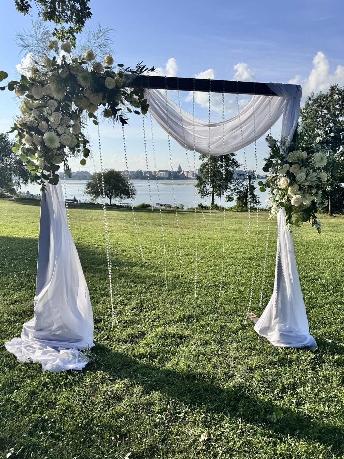 Outdoor wedding arch beside a lake with white draping, flowers and greenery