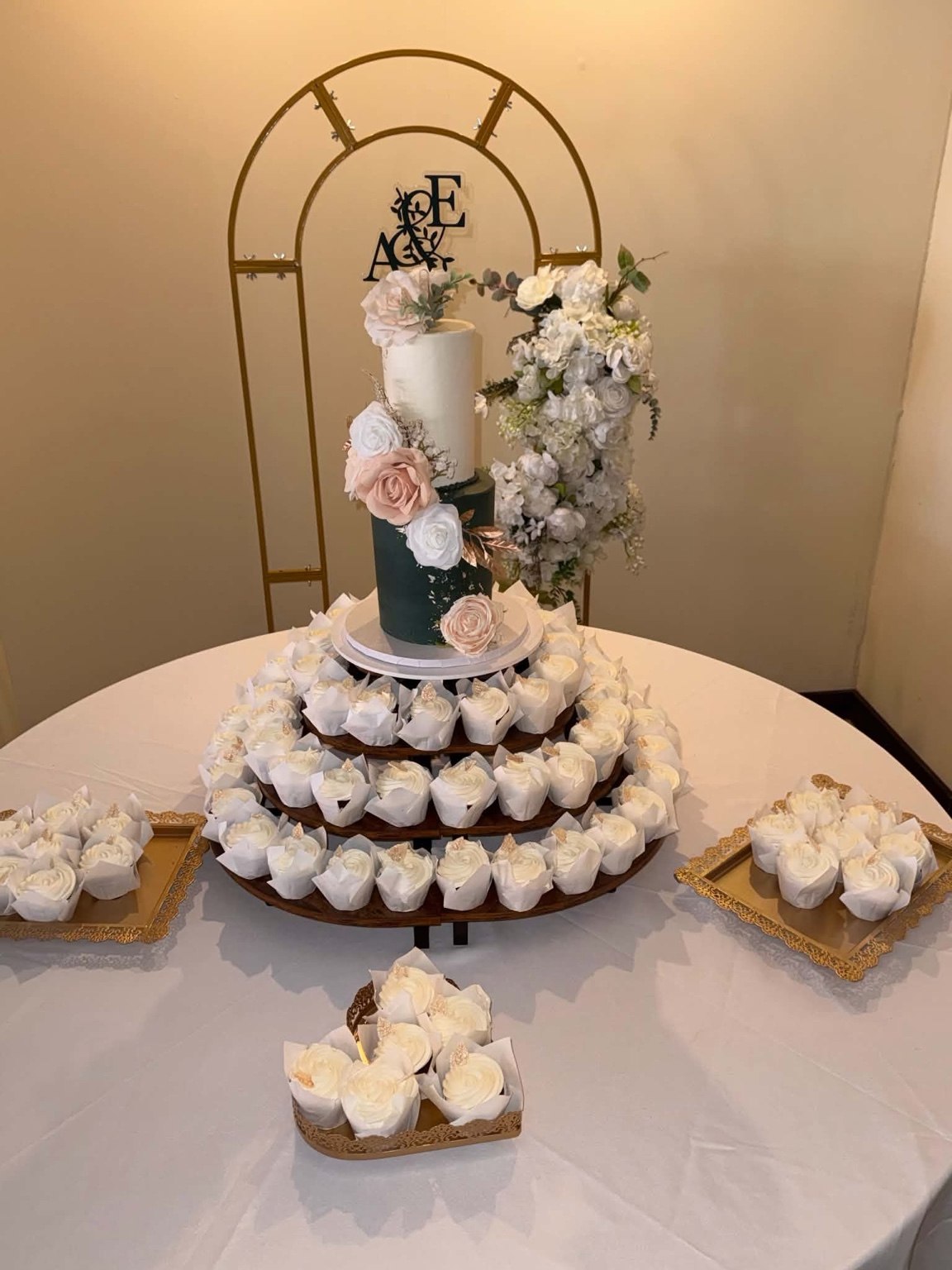 Tiered wedding cake with floral accents, cupcakes and a gold arch