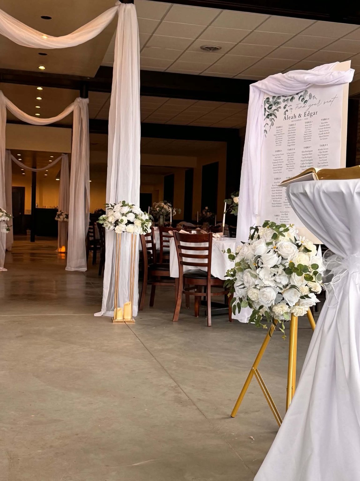 White draped entrance with white flowers on gold stands and a seating chart