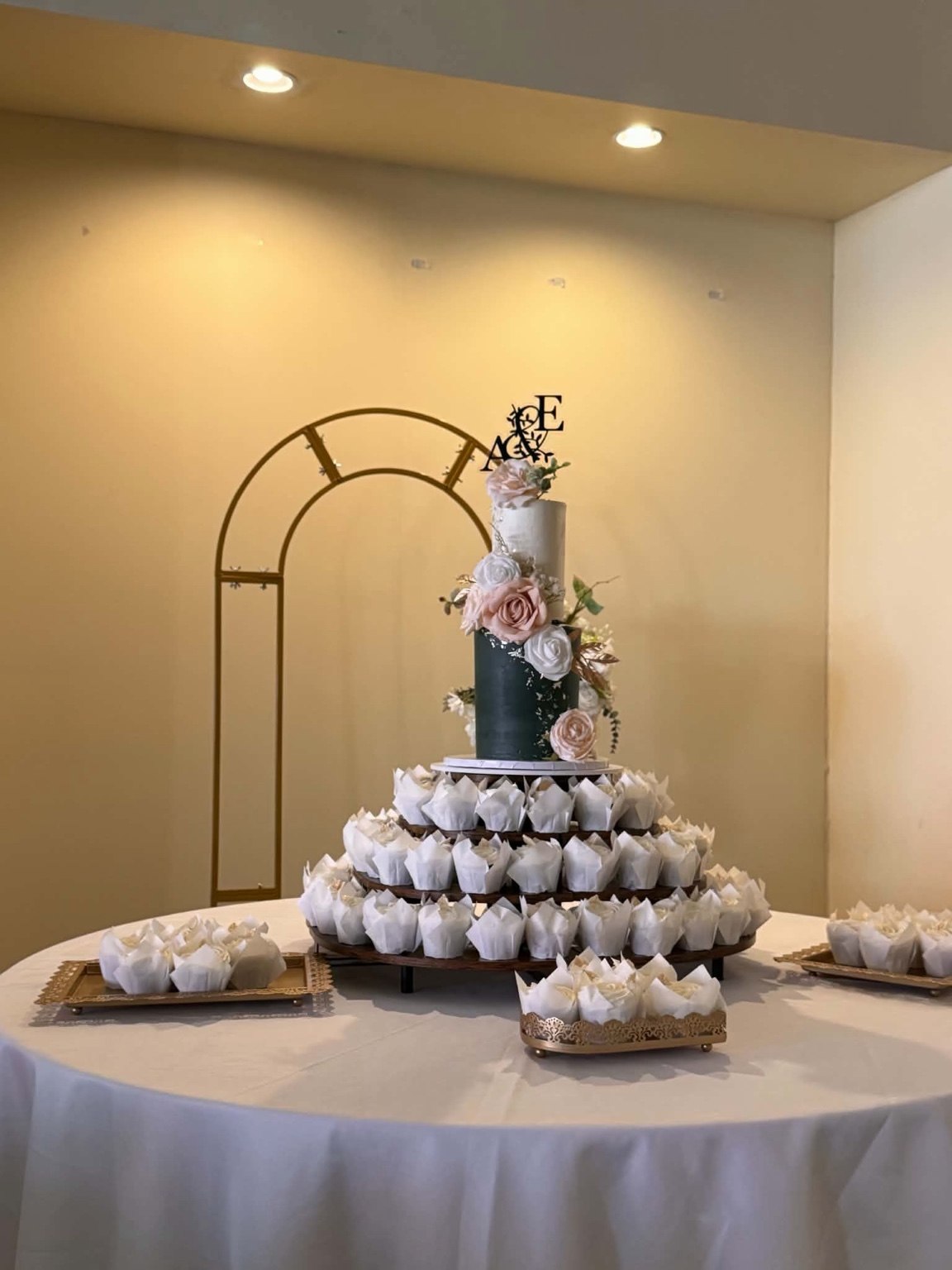 Wedding cake and cupcakes displayed in front of a gold arch