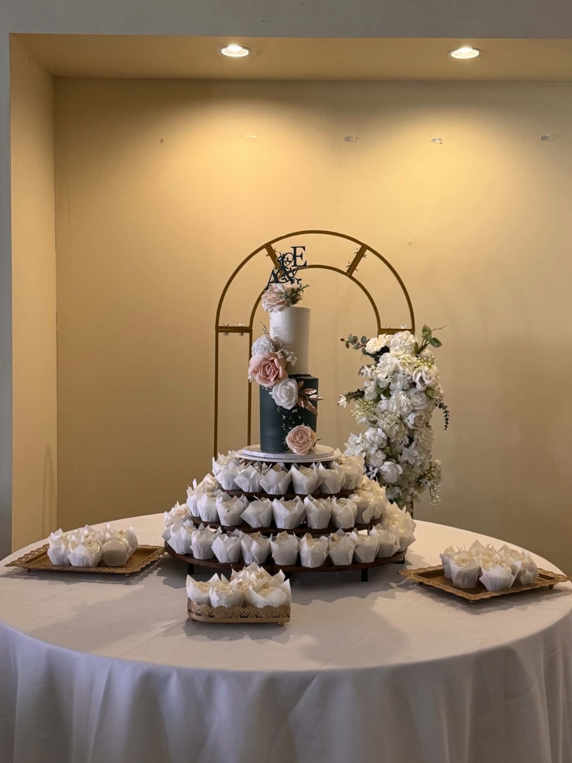 Wedding dessert display with a floral cake, cupcakes and gold arch